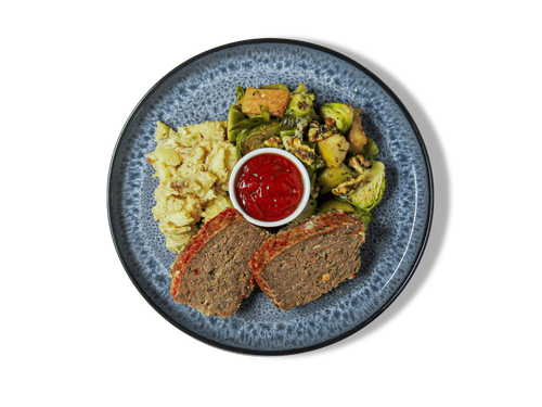 A top-down view of a plated meal on a white background, featuring two slices of meatloaf, Brussels sprouts, mashed potatoes, and a small bowl of red dipping sauce on a speckled blue-gray ceramic plate.